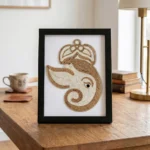 A framed artistic rope craft portrait of Lord Ganesha standing vertically on a polished wooden desk, styled next to stacked books, a coffee cup, and a desk lamp.