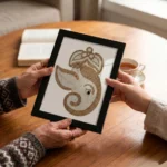 Two people holding a framed artistic jute rope craft portrait of Lord Ganesha over a wooden table, with an open book and a cup of tea in a warm indoor setting.