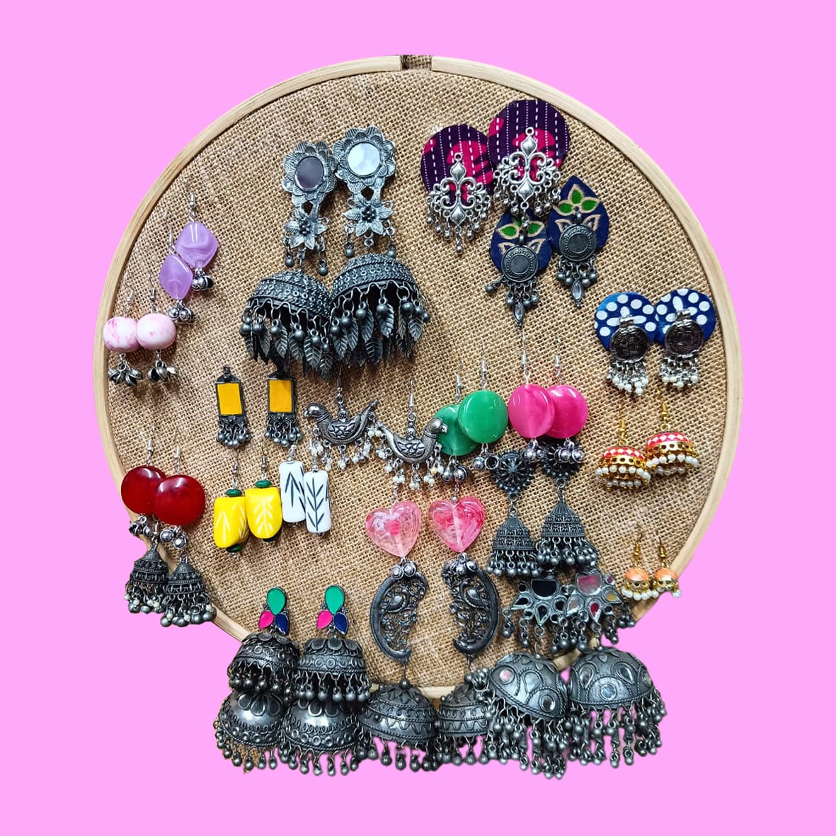 A display of handcrafted earrings including oxidized, colorful, and ethnic designs arranged on a round jute frame against a pink background.