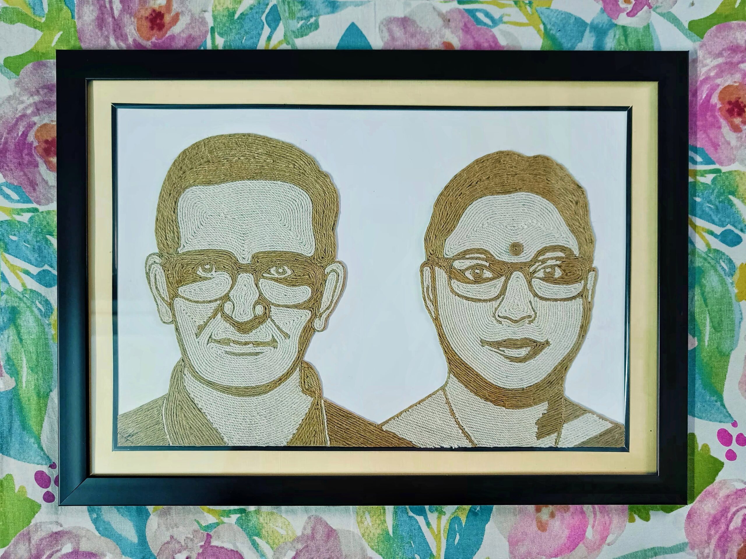 Framed handcrafted double portrait made entirely with jute rope, depicting an elderly man and woman wearing glasses, placed against a floral backdrop.