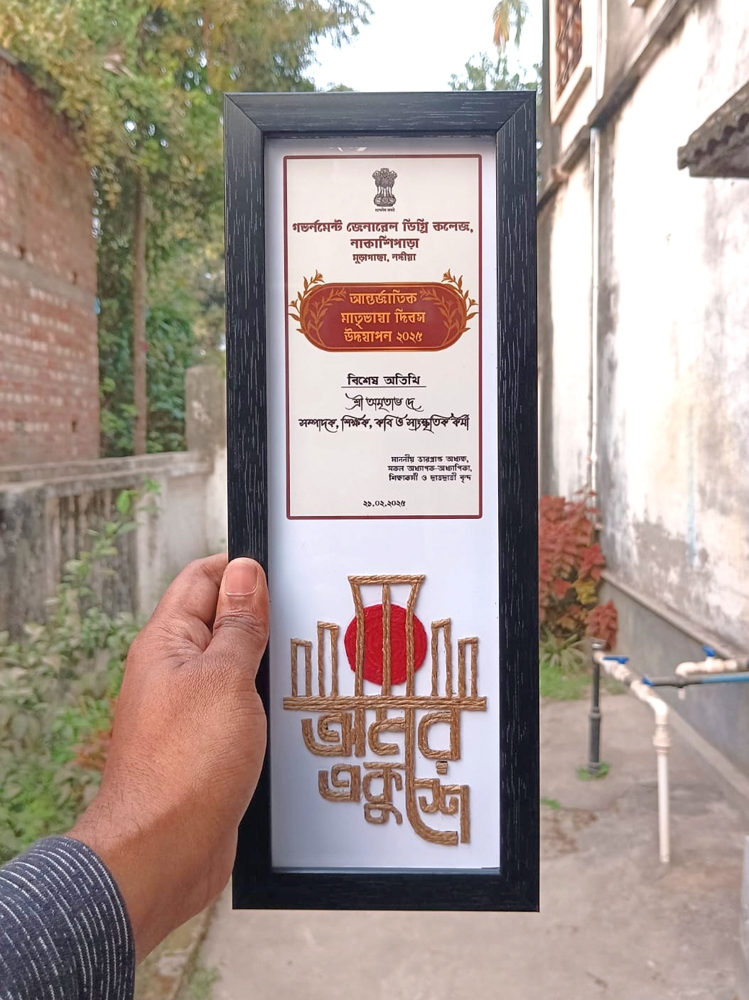Hand holding a handcrafted vertical frame featuring a Bengali invitation card for International Mother Language Day 2024 and a rope art of the Shahid Minar with "অমর একুশে" written in jute thread.