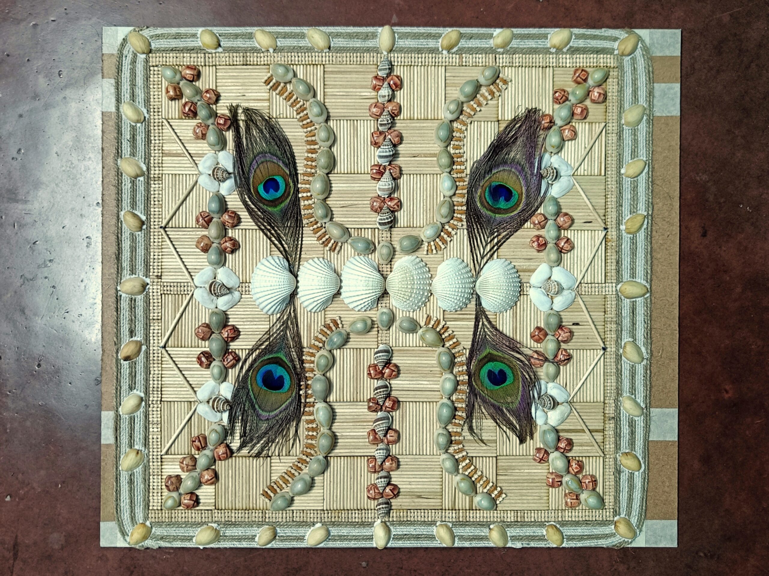 Intricate handmade wall hanging and card combo made with seashells, mustard seeds, tiny pods, and four peacock feathers arranged symmetrically on a bamboo mat base.