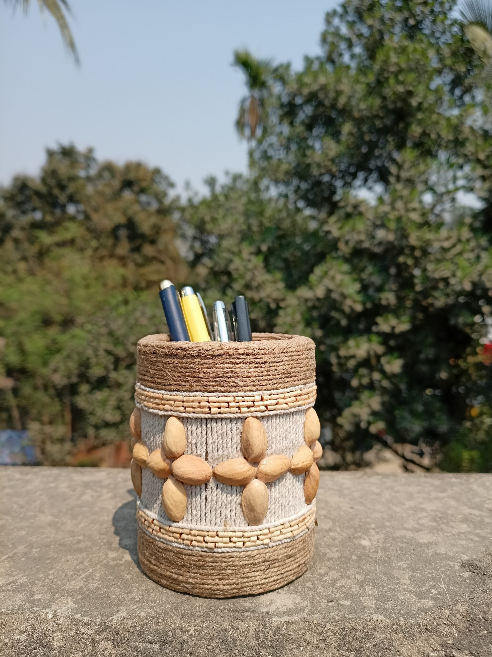 Handmade rustic pen stand decorated with natural jute rope, white threadwork, and pistachio shells arranged in floral patterns, placed outdoors with pens inside.