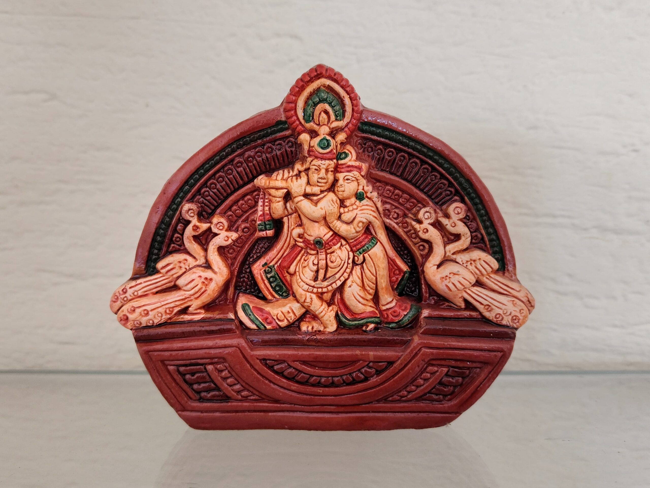 Handcrafted terracotta Radha-Krishna idol with peacocks on both sides, painted in earthy red, green, and beige tones, displayed against a plain backdrop.