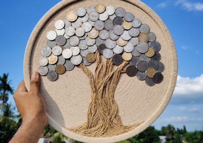 Hand holding a round handmade coin tree wall art against a blue sky background, made with real Indian coins and jute rope by Upahar18