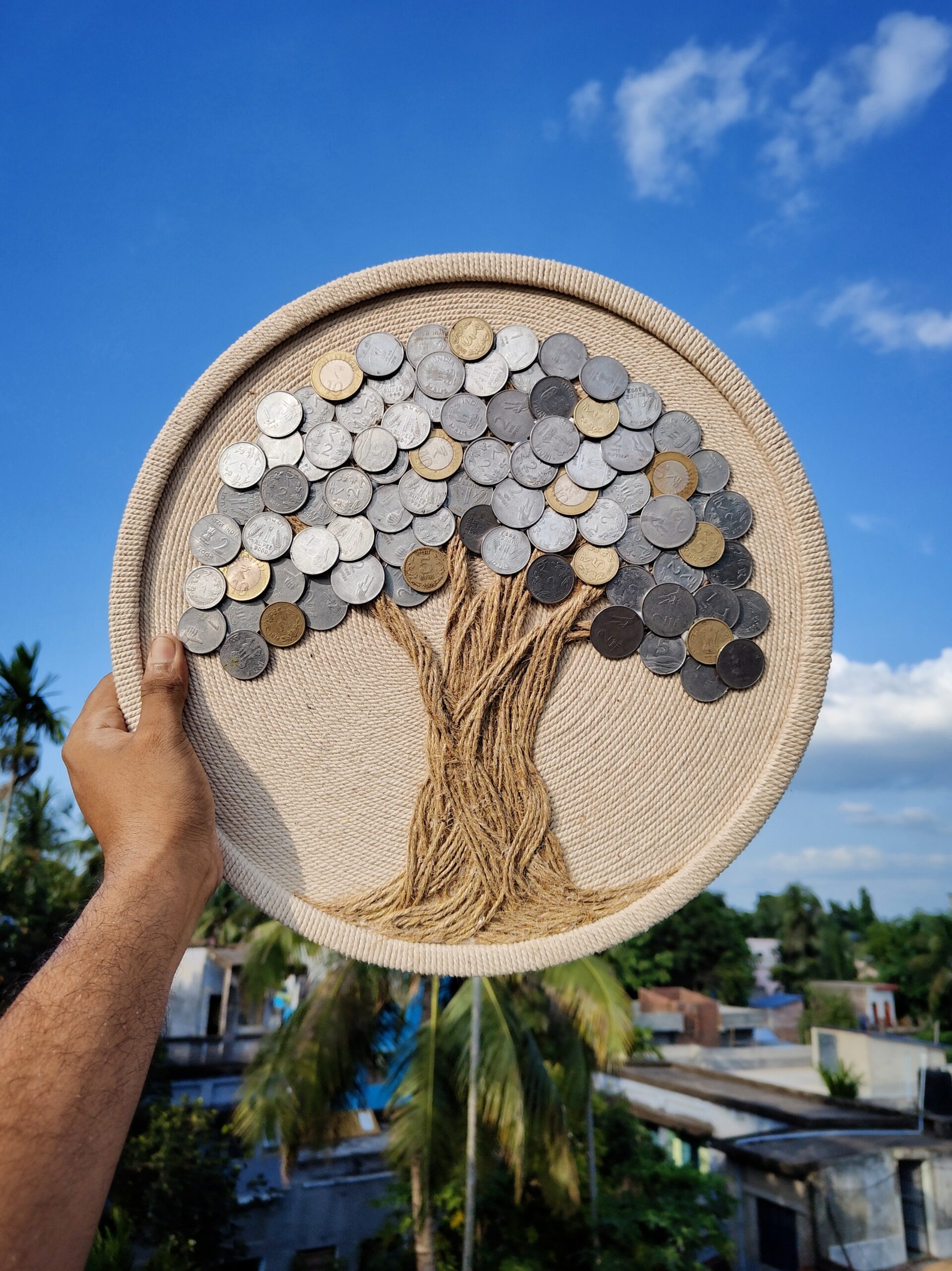 Hand holding a round handmade coin tree wall art against a blue sky background, made with real Indian coins and jute rope by Upahar18
