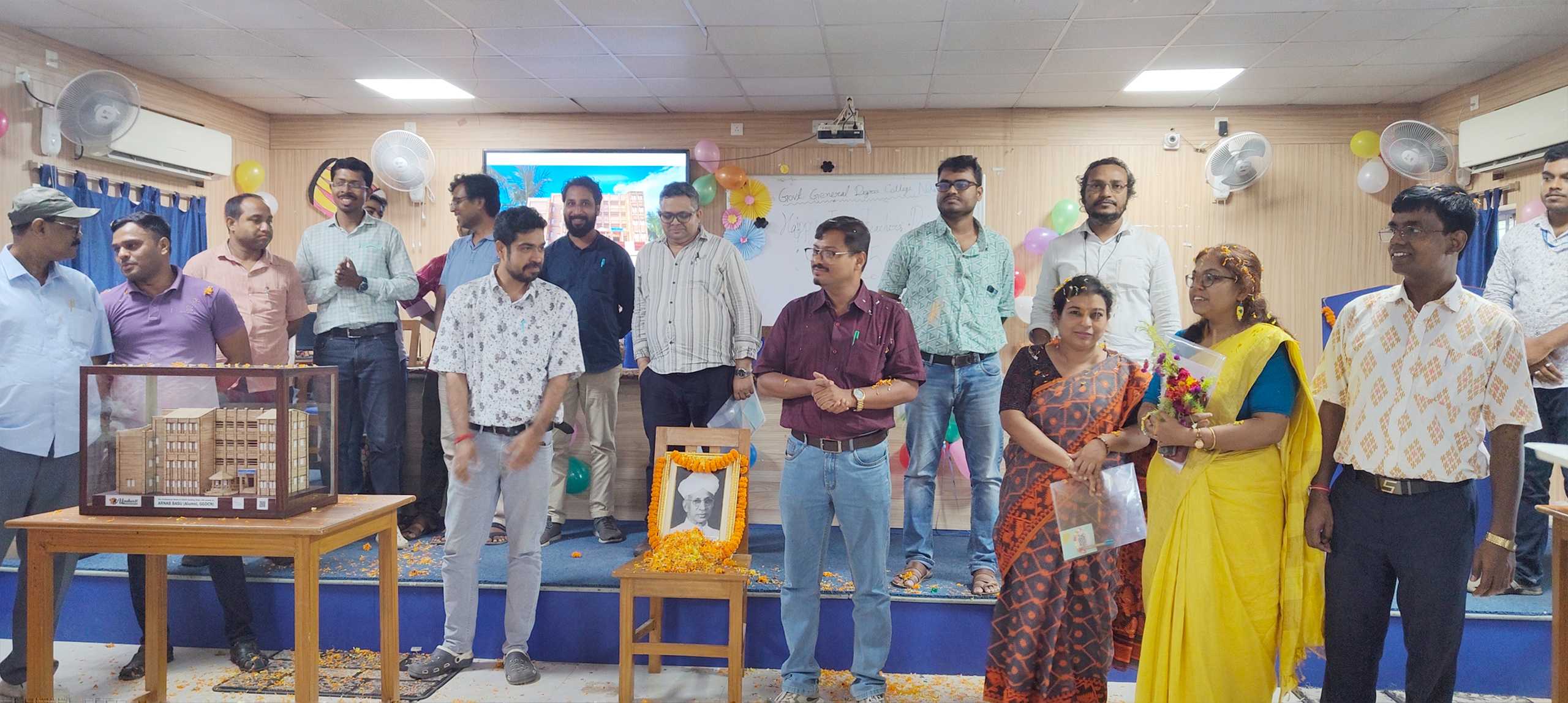 Professors and staff members of the college stand together on stage during the Teachers’ Day 2025 celebration, with the handcrafted architectural model of the college displayed inside a glass box on a wooden table and a decorated portrait of Dr. Sarvepalli Radhakrishnan placed nearby.