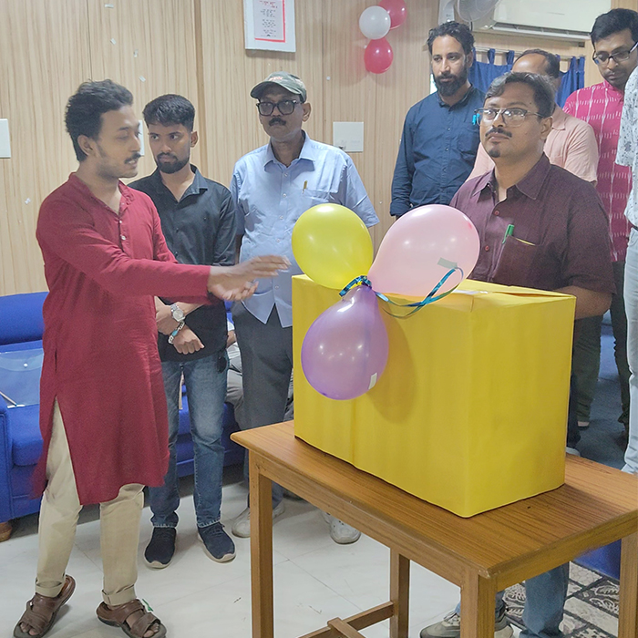 A group of people gathered indoors during a Teachers’ Day program. One person in a red kurta is standing beside a large yellow gift box decorated with balloons, while another person in a maroon shirt stands next to the box. Others are standing in the background observing.