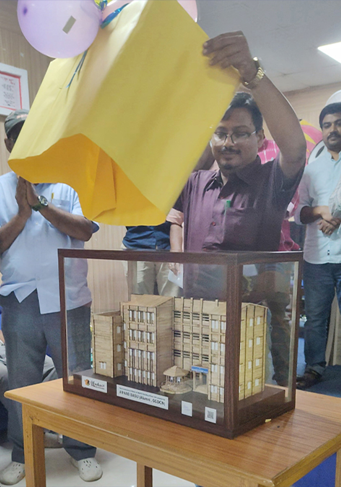 The Officer-in-Charge is unveiling a handmade architectural model of the college by lifting a yellow cover, while others stand nearby and watch with admiration.