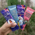 A hand holding four customized Cadbury chocolate bars with personalized photos and romantic or affectionate Bengali typography printed on differently colored wrappers against a blurry green background.