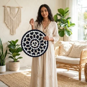 A woman holding the Cowrie and Seashell Mandala Handcrafted Round Wall Decor by Upahar18 to showcase its actual size and scale in a brightly lit room.