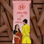 A pre-designed soft pink chocolate wrapper featuring the famous lyrical Bengali Quote: "Batase Bohiche Prem Noyone Lagilo Nesha." This Romantic themed KitKat design illustrates a couple where the man in a yellow panjabi is hiding a rose behind his back for the woman in a gorgeous saree, capturing a sweet surprise moment.
