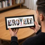 An over-the-shoulder view of a person holding a classic black rectangular Upahar18 Brother Photo Frame displaying a custom-printed layout of the word BROTHER filled with a photo collage, featuring World's Best on top and Happy Raksha Bandhan at the bottom.