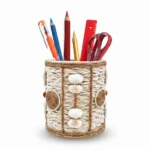 Studio photograph against a white background of a handmade rope pen stand with white seashells and brown twine circles, containing colorful pencils, pens, a red ruler, and red scissors.