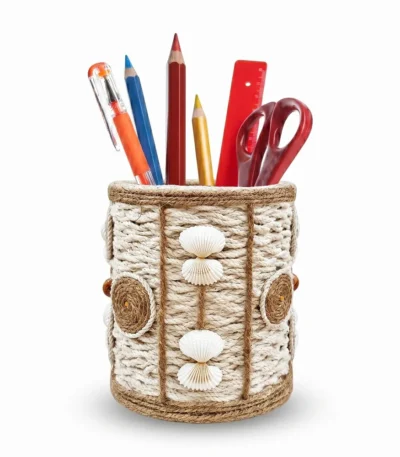 Studio photograph against a white background of a handmade rope pen stand with white seashells and brown twine circles, containing colorful pencils, pens, a red ruler, and red scissors.