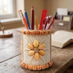 Upahar18 Handmade Eco-Friendly Rope Base Pen Stand on a rustic wooden study desk near a window, showcasing beautiful mirror work and red bead floral patterns. This artistic desk organiser holds various stationery items like pens, pencils, a scale, and scissors.