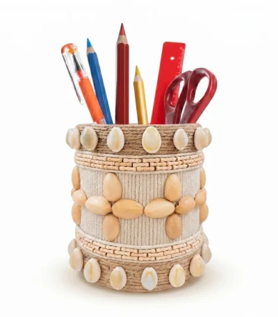 Studio shot of Upahar18 Handmade Eco-Friendly Rope Base Pen Stand with Cowrie Shell borders against a white background. This artisanal desk organiser is filled with pencils, pens, and scissors, highlighting the polished cowrie shells lining the top and bottom rims along with a central wooden seed flower pattern.