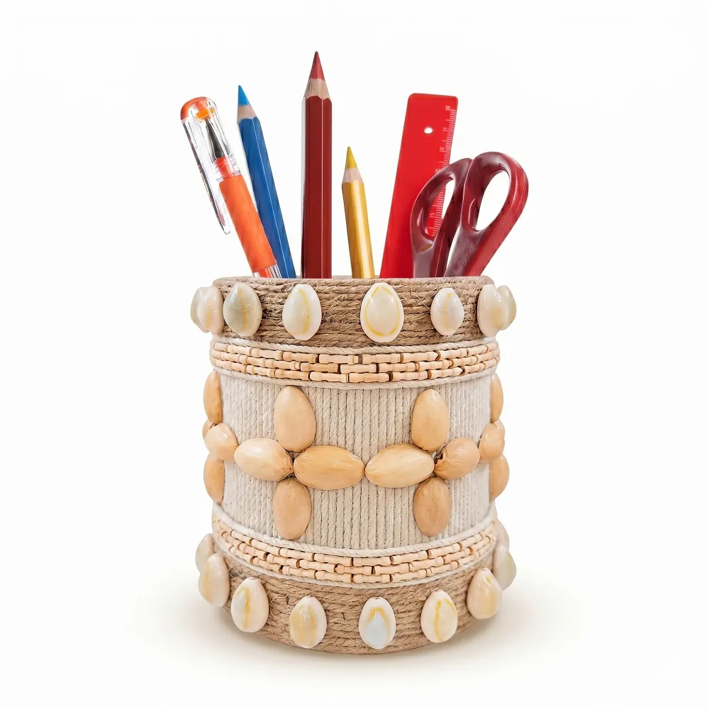 Studio shot of Upahar18 Handmade Eco-Friendly Rope Base Pen Stand with Cowrie Shell borders against a white background. This artisanal desk organiser is filled with pencils, pens, and scissors, highlighting the polished cowrie shells lining the top and bottom rims along with a central wooden seed flower pattern.