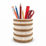 Studio shot of Upahar18 Handmade Eco-Friendly Rope Base Pen Stand against a white background. This minimal desk organiser features a cream-colored woven rope cylinder encircled by five horizontal dark brown jute rope bands, holding colorful pencils, pens, a scale, and scissors.