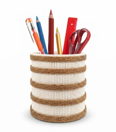 Studio shot of Upahar18 Handmade Eco-Friendly Rope Base Pen Stand against a white background. This minimal desk organiser features a cream-colored woven rope cylinder encircled by five horizontal dark brown jute rope bands, holding colorful pencils, pens, a scale, and scissors.