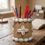 Upahar18 Handmade Eco-Friendly Rope Base Pen Stand neatly arranged on a rustic wooden study desk near an open book. The brown jute and white seashell desk organiser holds vibrant writing tools, creating a bohemian and coastal aesthetic in a sunlit room.