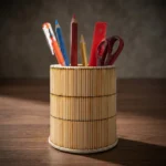 A cylindrical handmade pen stand on a dark wooden office desk, creatively designed with vertical multicolored wooden sticks. The holder contains colorful pencils, a black pen, a red ruler, and scissors, set against a warm, softly blurred study room background.
