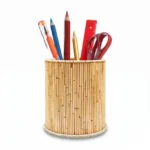 An isolated product shot of a round handmade pen stand by Upahar18 on a solid white background. The holder is covered tightly in slender, light brown natural wooden sticks and is filled with essential stationery items like pencils, a pen, and office scissors.