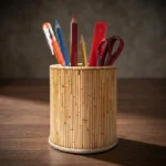 A natural tan cylindrical handmade pen stand resting on a dark wooden office desk. Crafted with a texture of thin vertical natural wooden sticks, it holds stationery including an orange pen, color pencils, a red ruler, and scissors under warm, artistic office lighting.