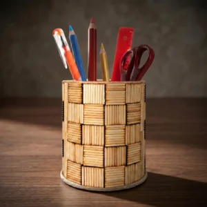 A cylindrical handmade pen stand featuring an artistic checkerboard pattern made from blocks of tiny natural wooden sticks, placed on a warm dark wooden desk. The organizer holds stationery like pencils, an orange pen, a red ruler, and maroon scissors under soft studio lighting.