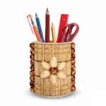 An isolated high-angle product shot of the Upahar18 handmade stationery holder against a white backdrop. The round stand is adorned with thin natural wooden sticks, side borders of dark brown glossy beads, and a central flower motif made from cream-colored pistachio shells, holding basic office stationery.