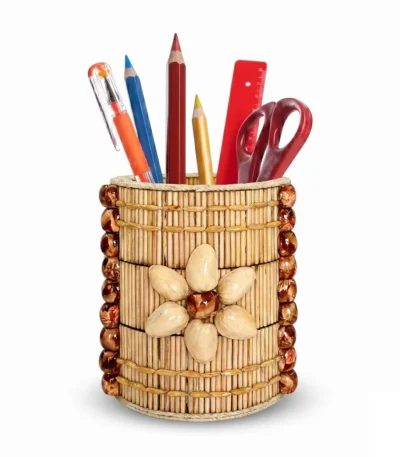 An isolated high-angle product shot of the Upahar18 handmade stationery holder against a white backdrop. The round stand is adorned with thin natural wooden sticks, side borders of dark brown glossy beads, and a central flower motif made from cream-colored pistachio shells, holding basic office stationery.