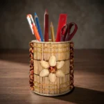 A cylindrical handmade pen stand on a dark wooden office desk under warm studio lighting. The holder is uniquely decorated with vertical natural wooden sticks, vertical rows of brown polished beads on the sides, and a prominent 6-petaled flower crafted from natural pistachio shells at the center. It holds colorful pencils, an orange pen, a red ruler, and scissors.