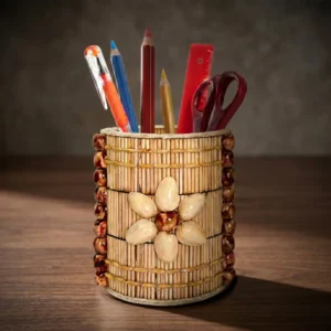 A cylindrical handmade pen stand on a dark wooden office desk under warm studio lighting. The holder is uniquely decorated with vertical natural wooden sticks, vertical rows of brown polished beads on the sides, and a prominent 6-petaled flower crafted from natural pistachio shells at the center. It holds colorful pencils, an orange pen, a red ruler, and scissors.