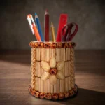 A cylindrical handmade pen holder decorated with a central pistachio shell flower and rich brown bead borders, resting on a polished dark wooden office desk. Photographed under dramatic warm studio lighting, the container holds daily office stationery against a soft-focus background.