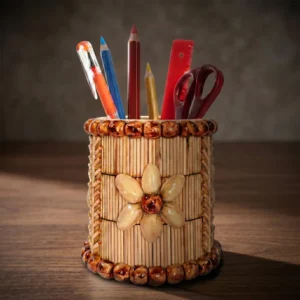 A cylindrical handmade pen holder decorated with a central pistachio shell flower and rich brown bead borders, resting on a polished dark wooden office desk. Photographed under dramatic warm studio lighting, the container holds daily office stationery against a soft-focus background.