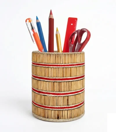 A studio shot of the Upahar18 handmade pen stand against a solid white background. The cylindrical container is crafted with closely packed vertical natural wooden sticks, accented by horizontal red and cream striped bands wrapping around the body. It holds colorful pencils, a red ruler, a pen, and office scissors.
