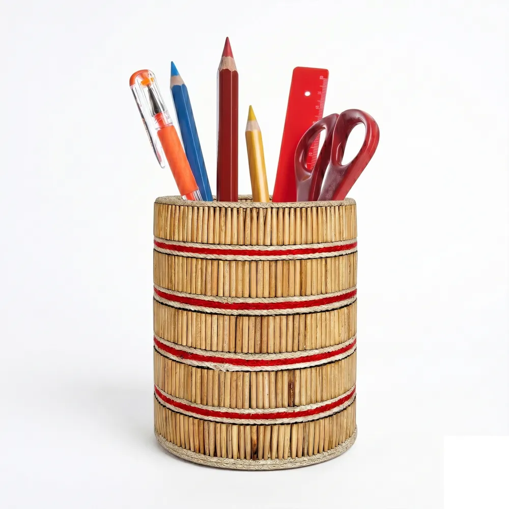 A studio shot of the Upahar18 handmade pen stand against a solid white background. The cylindrical container is crafted with closely packed vertical natural wooden sticks, accented by horizontal red and cream striped bands wrapping around the body. It holds colorful pencils, a red ruler, a pen, and office scissors.