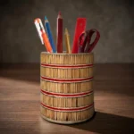 A cylindrical handmade pen holder with natural wood textures and red horizontal stripes, resting on a polished dark wooden office desk. Photographed under warm, artistic studio lighting, the stand is filled with stationery items against a soft-focus blurred background.