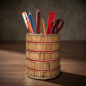 A cylindrical handmade pen holder with natural wood textures and red horizontal stripes, resting on a polished dark wooden office desk. Photographed under warm, artistic studio lighting, the stand is filled with stationery items against a soft-focus blurred background.