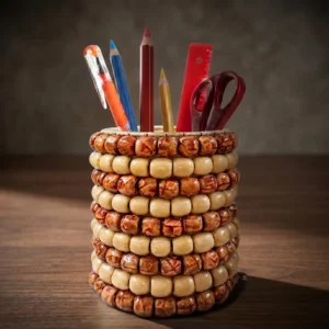 A cylindrical handmade pen stand crafted from alternating rows of dark patterned and light cream wooden beads, placed on a dark wooden desk. Inside the holder are colorful pencils, a pen, a red ruler, and a pair of scissors, creating a professional office desk setup with a soft-focus background.