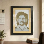 A framed rope craft portrait of MS Dhoni hanging on a beige office wall beside a calendar, overlooking a desk with books and an indoor green plant.