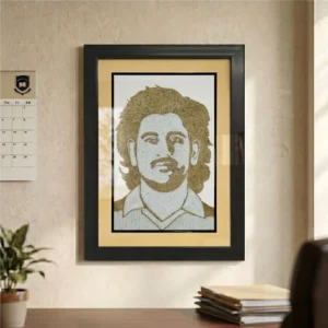 A framed rope craft portrait of MS Dhoni hanging on a beige office wall beside a calendar, overlooking a desk with books and an indoor green plant.