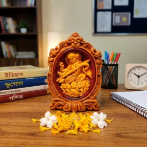 Handcrafted upahar18 Terracotta Maa Saraswati idol displayed on a study table next to notebooks, text books, and pen stand with loose marigold flowers, made in Krishnanagar.