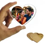 A person holding a heart-shaped clear acrylic magnetic fridge magnet frame showing a personalized photo of a smiling Indian couple in traditional wedding attire, with a protective brown kraft paper film lying on a white background by upahar18.