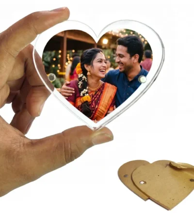 A person holding a heart-shaped clear acrylic magnetic fridge magnet frame showing a personalized photo of a smiling Indian couple in traditional wedding attire, with a protective brown kraft paper film lying on a white background by upahar18.