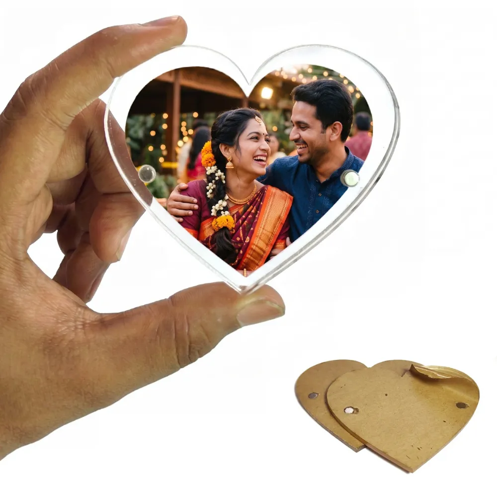 A person holding a heart-shaped clear acrylic magnetic fridge magnet frame showing a personalized photo of a smiling Indian couple in traditional wedding attire, with a protective brown kraft paper film lying on a white background by upahar18.