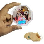 A hand holding an eight-sided octagon-shaped transparent acrylic magnetic photo frame with a customized family portrait inside, displaying an unpeeled brown protective paper sheet underneath on a white surface by upahar18.