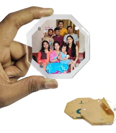 A hand holding an eight-sided octagon-shaped transparent acrylic magnetic photo frame with a customized family portrait inside, displaying an unpeeled brown protective paper sheet underneath on a white surface by upahar18.