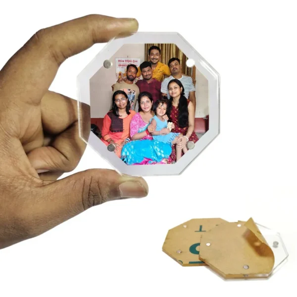 A hand holding an eight-sided octagon-shaped transparent acrylic magnetic photo frame with a customized family portrait inside, displaying an unpeeled brown protective paper sheet underneath on a white surface by upahar18.