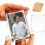 A close-up view of a hand holding a vertical rectangle-shaped personalized acrylic magnetic fridge magnet frame containing a photo of a man in sunglasses, with a separate blank clear block and a brown paper covered piece in the background by upahar18.
